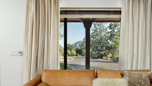A room with a brown leather sofa, a white fluffy pillow, and a patterned pillow. A window with beige curtains reveals a view of trees and a mountain.