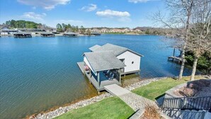 A lakeside dock with a small cabin, clear water, and a distant view of houses and trees. - Gentle Slope Lakefront beautiful sunset,6Bds,4.5Bas, kayaks,paddleboards,firepit (Moneta)