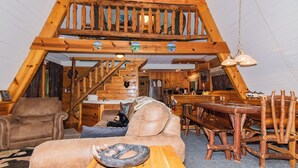 Interior acogedor de una cabaña de madera con un dormitorio en el altillo, un comedor y una sala de estar amueblada con sofás de cuero y mesas de madera.