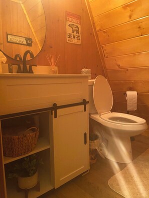 Ein kleines Badezimmer mit Toilette, einem kleinen Waschbecken und einem Spiegel. An der Wand befindet sich ein Schild und ein Korb unter dem Waschbecken.