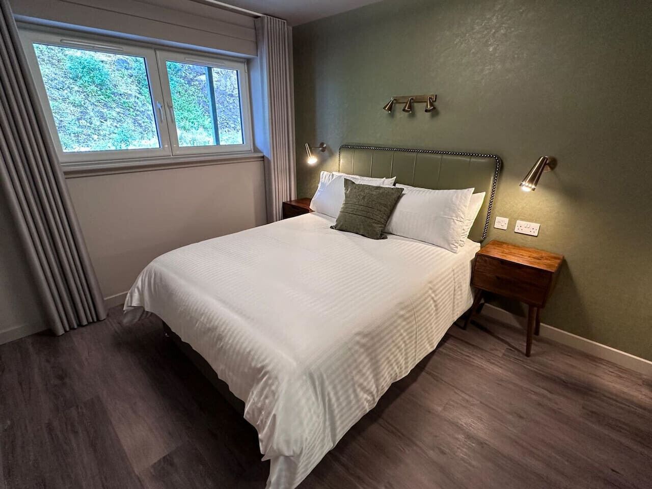 Una cama bien tendida con sábanas blancas, cabecera con luces empotradas en la pared, mesita de noche de madera y una ventana con cortinas.