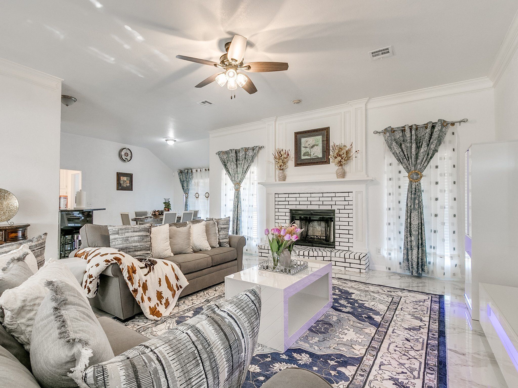 A spacious living room with a fireplace, a ceiling fan, and a dining area in the background.