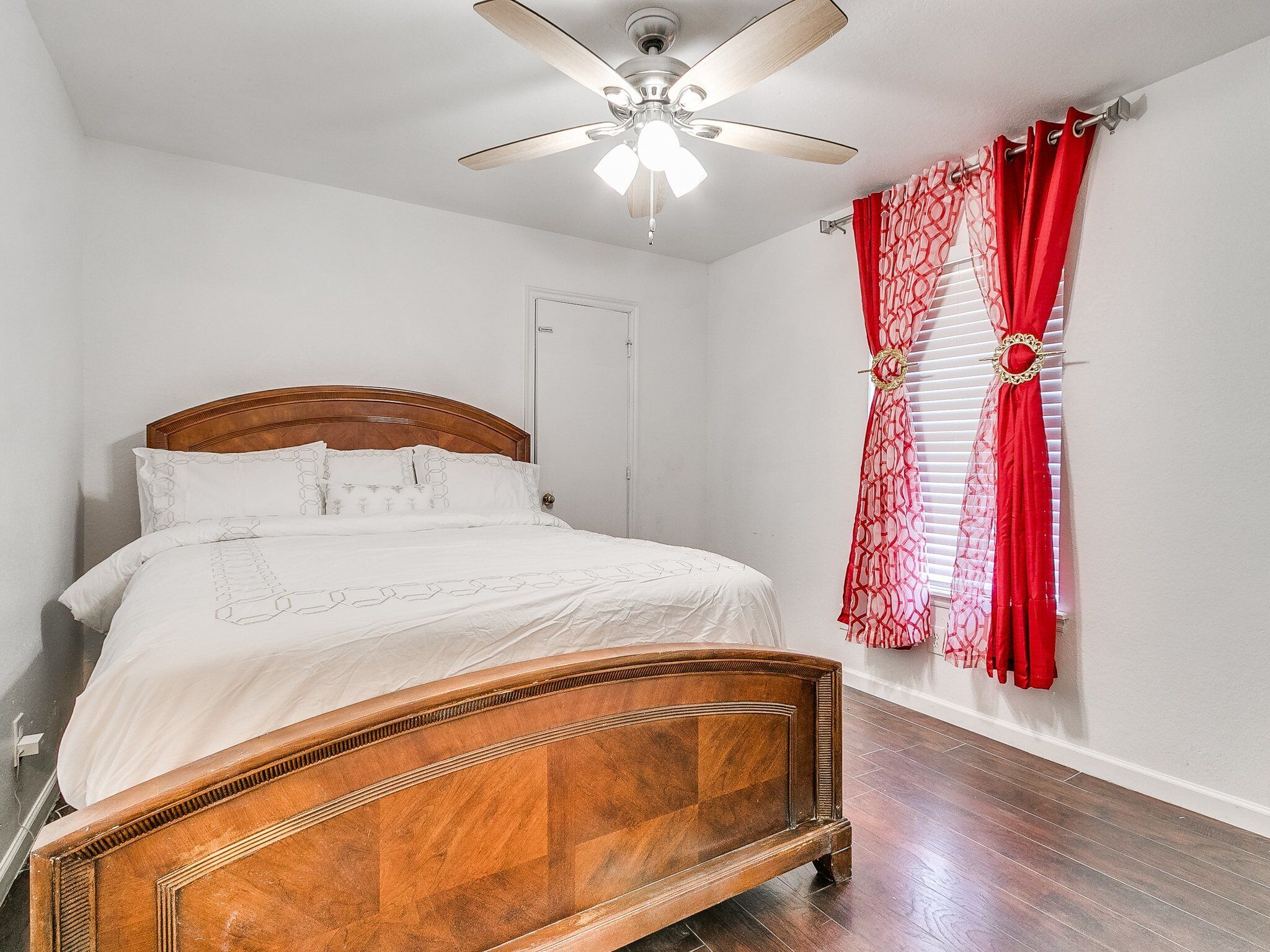 A bedroom with a wooden bed, white bedding, a ceiling fan, and red patterned curtains.