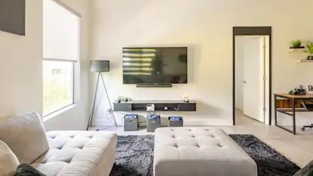 Ein modernes Wohnzimmer mit großem Flachbildfernseher an der Wand, einer Couch, einem Hocker und einem kleinen Beistelltisch.