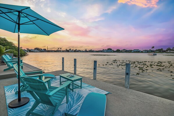 A waterfront patio with teal lounge chairs and a table, overlooking a calm body of water and a sunset sky.