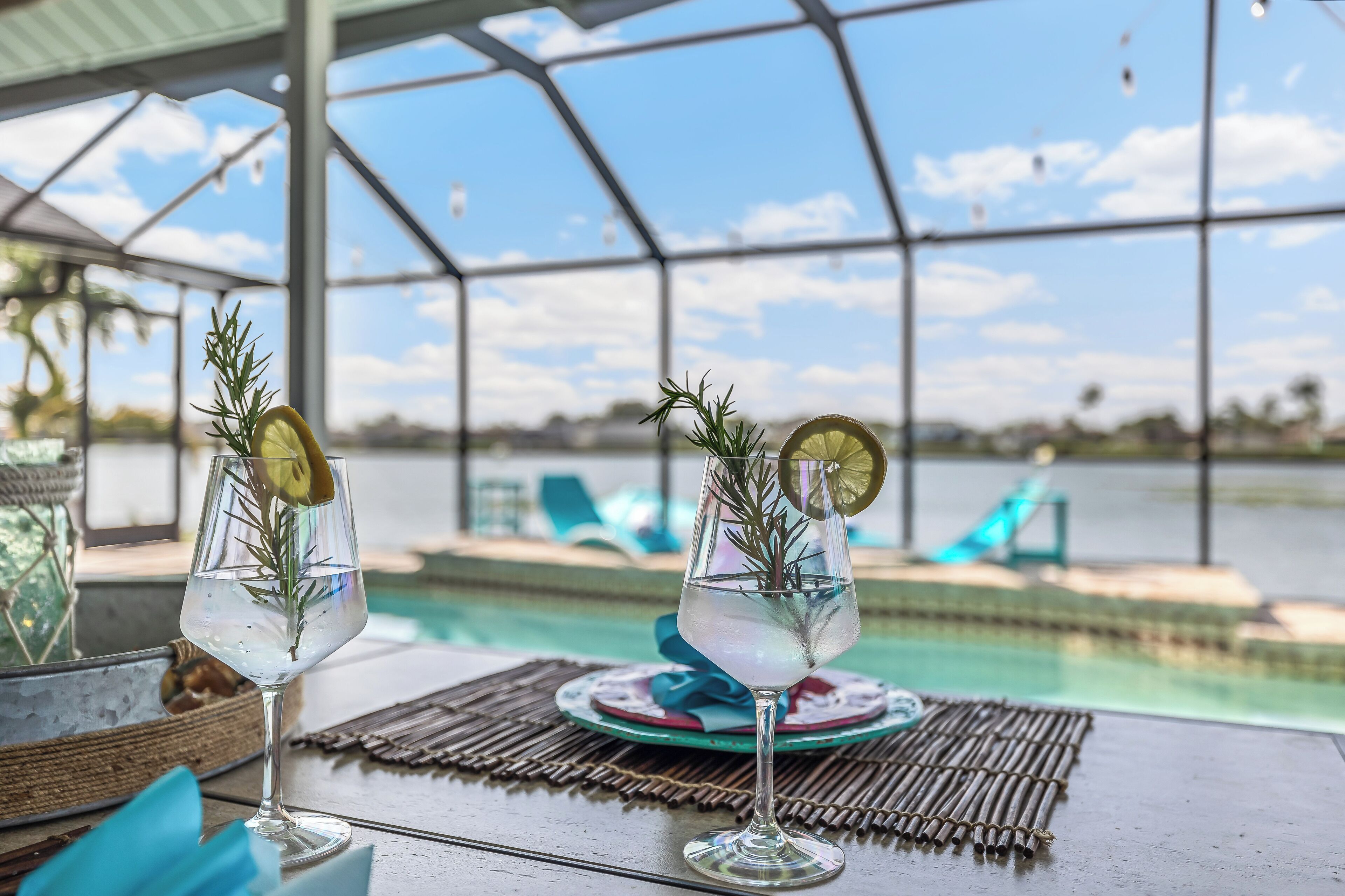 Two glasses of a clear beverage with rosemary and a lime slice, placed on a table with a view of a pool and lounge chairs.