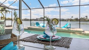 To glas af en klar drik med rosmarin og et limeskive, placeret på et bord med udsigt til en pool og liggestole.