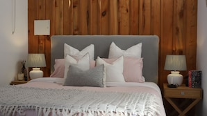 A neatly made bed with a grey headboard, pink and white pillows, and a white throw blanket. There are two bedside tables with lamps and books. - Barn Stay Hideaway "The Barn" (Somerville)