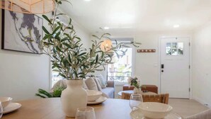 Un comedor con una mesa de madera dispuesta para cuatro personas, un jarrón blanco con follaje y un cuadro en la pared.
