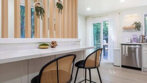 Una cocina moderna con encimera blanca, dos taburetes negros, listones de madera en la pared y un refrigerador de acero inoxidable.