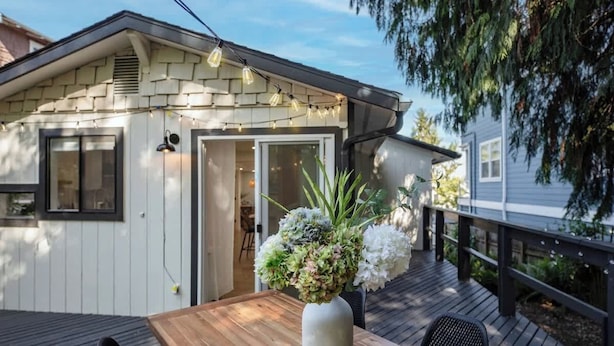 Una terraza de madera con una mesa y sillas, un muro exterior blanco y una puerta que da a un espacio interior.