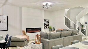 A modern living room with a grey sectional sofa, a dining table with chairs, a fireplace, and a staircase with a railing. - Livingston Gem: 5BR Home with Chef's Kitchen (Calgary)