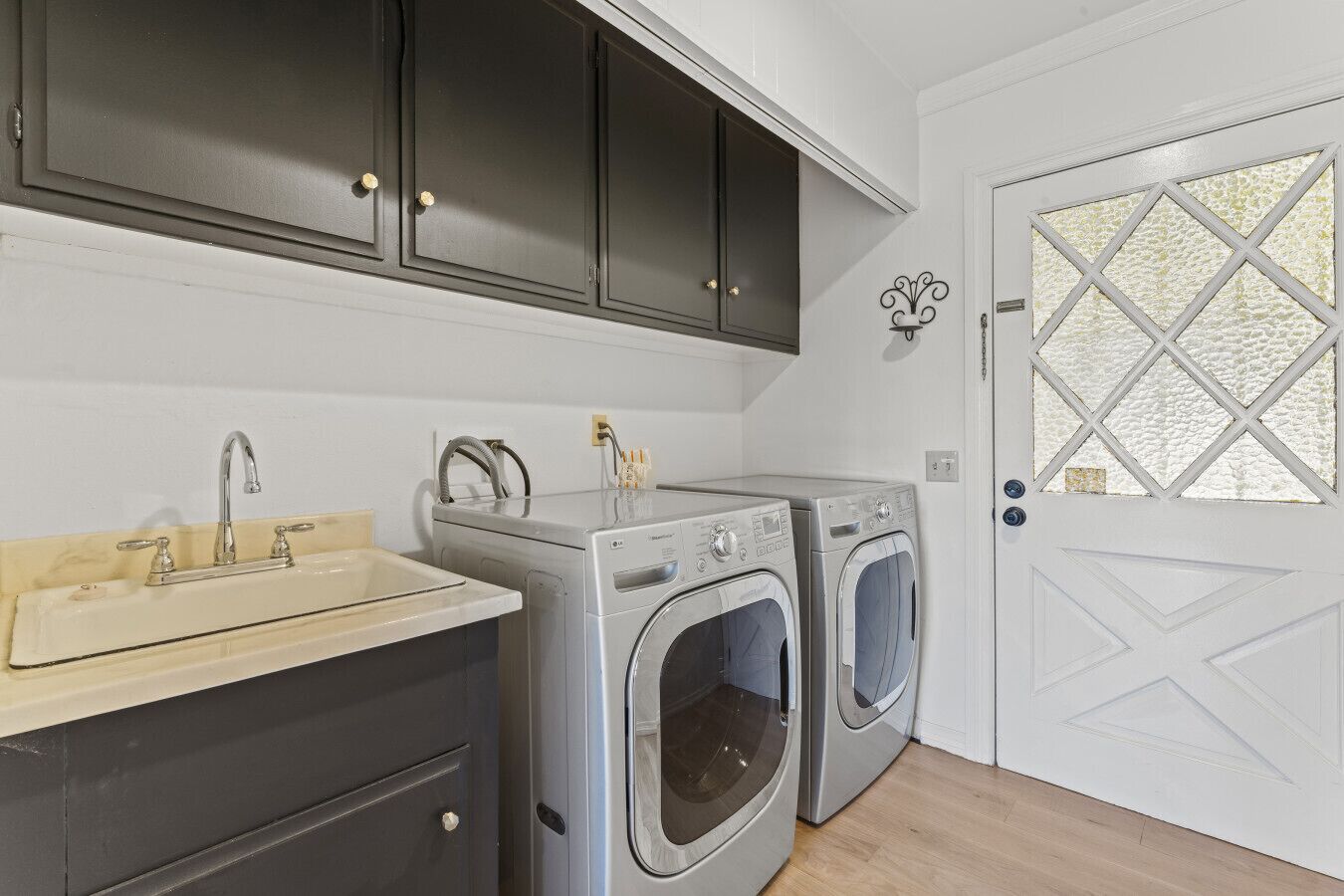 A compact laundry room with two front-loading washing machines, a sink with a faucet, and dark cabinets above.