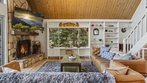 Una sala de estar acogedora con chimenea de piedra, un ventanal con vista a árboles, un sofá de cuero y un techo de madera.