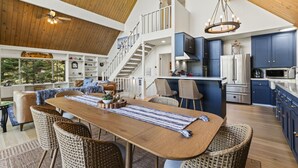 Amplia sala de estar con techo alto, una mesa de comedor de madera, sillas de mimbre, una escalera que lleva a un nivel superior y una cocina con armarios azules.