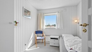 A small, white room with a bed, a chair, a desk, and a window with curtains.