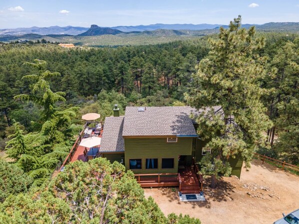 Una casa de dos pisos con terraza, rodeada por un bosque frondoso y montañas al fondo.
