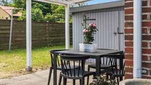 Un patio con una mesa y cuatro sillas, una planta en maceta con flores rosas, y un cobertizo de madera al fondo.