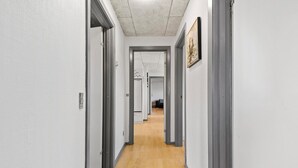 Un pasillo estrecho con paredes blancas y suelo de madera, que conduce a una habitación con una cama y una silla.