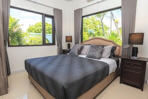 A bedroom with a large bed, two bedside tables, a headboard, and a view of greenery outside.