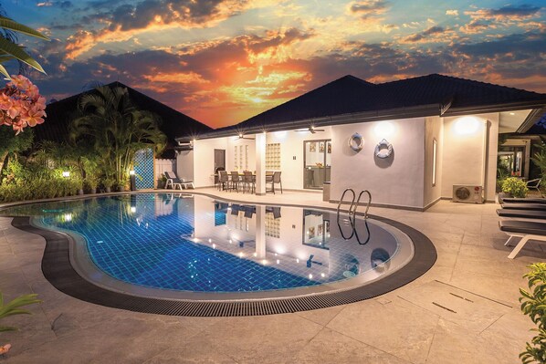 A swimming pool with a modern house in the background, a covered patio, and a sunset sky.