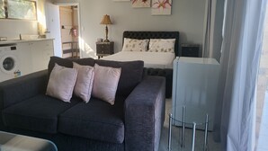 A hotel room with a dark grey sofa, a bed with white bedding, a small table with a lamp, and a TV stand. - Top View Deluxe Self-Contained Room No 3 with Balcony Overlooking Swimming Pool. (Windsor)