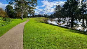 A park with a paved walkway, lush green grass, and a calm body of water reflecting trees. - Top View Deluxe Self-Contained Room No 3 with Balcony Overlooking Swimming Pool. (Windsor)