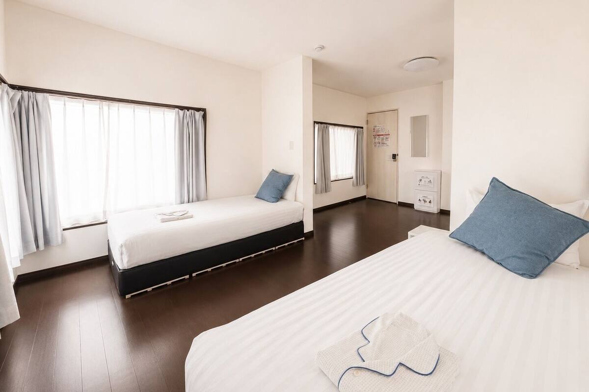 A hotel room with two beds, a large window with curtains, a door, and a small white cabinet.