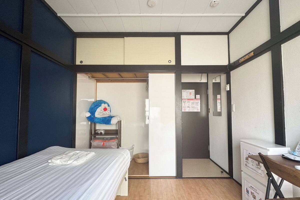 A room with a bed, a desk, a chair, a wardrobe, and a door leading to another room.