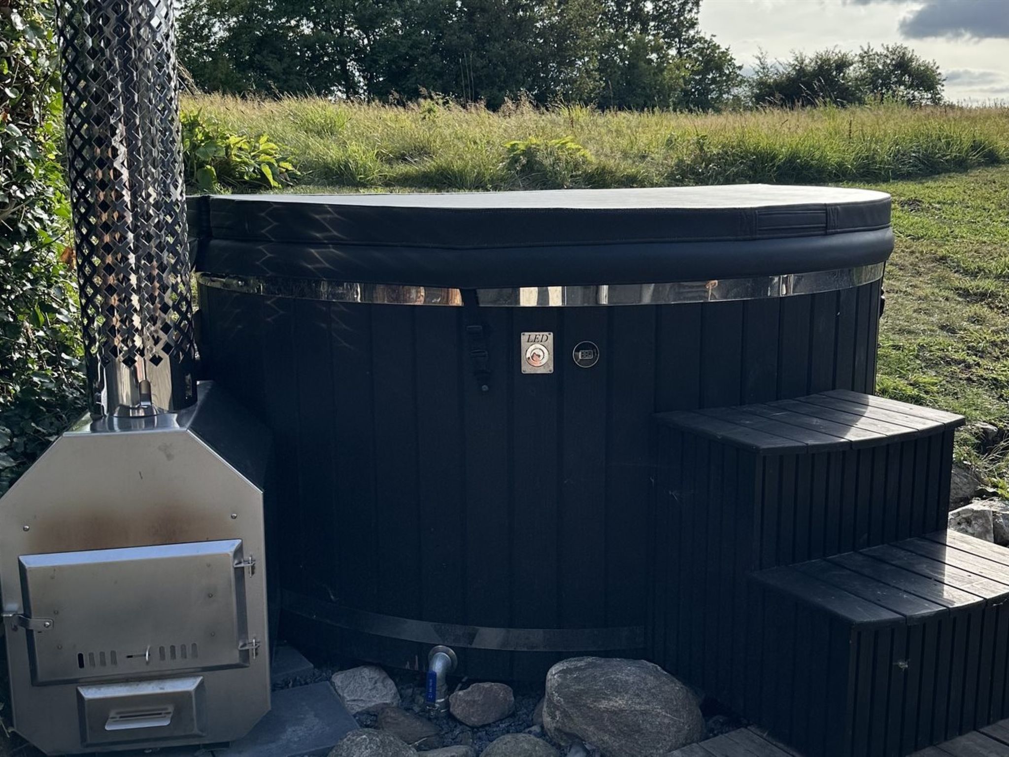 Un jacuzzi con estructura metálica y panel de control, rodeado de rocas y follaje.