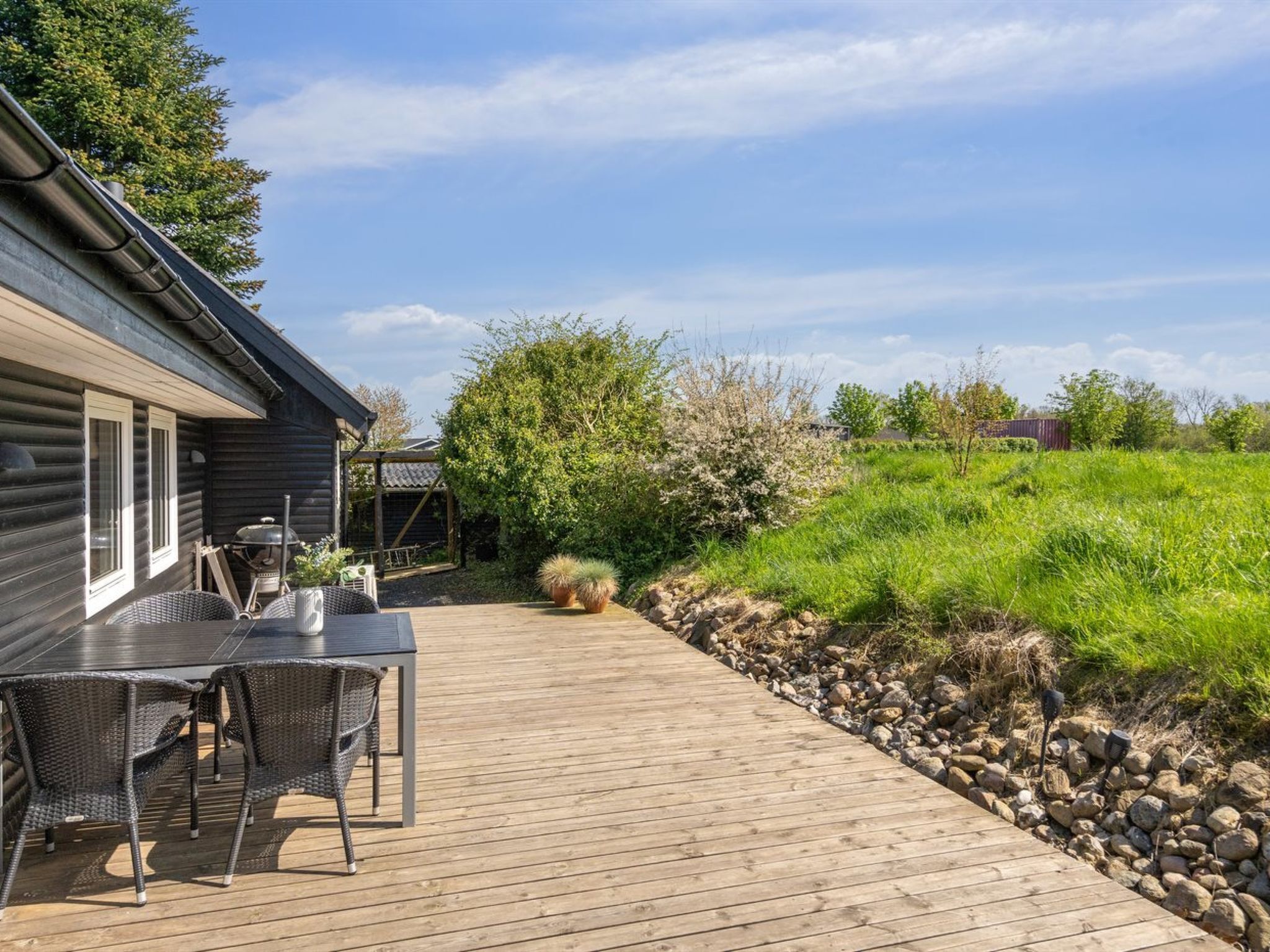 A wooden deck with a table and chairs, surrounded by greenery and a stone wall.