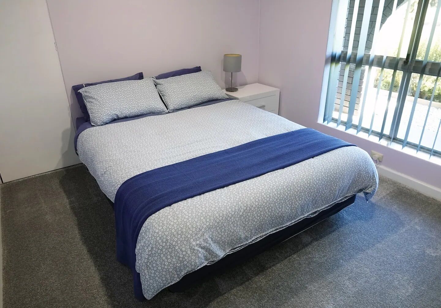 Un lit bien fait, avec une couette bleue et blanche, une lampe de chevet et une fenêtre avec des stores.