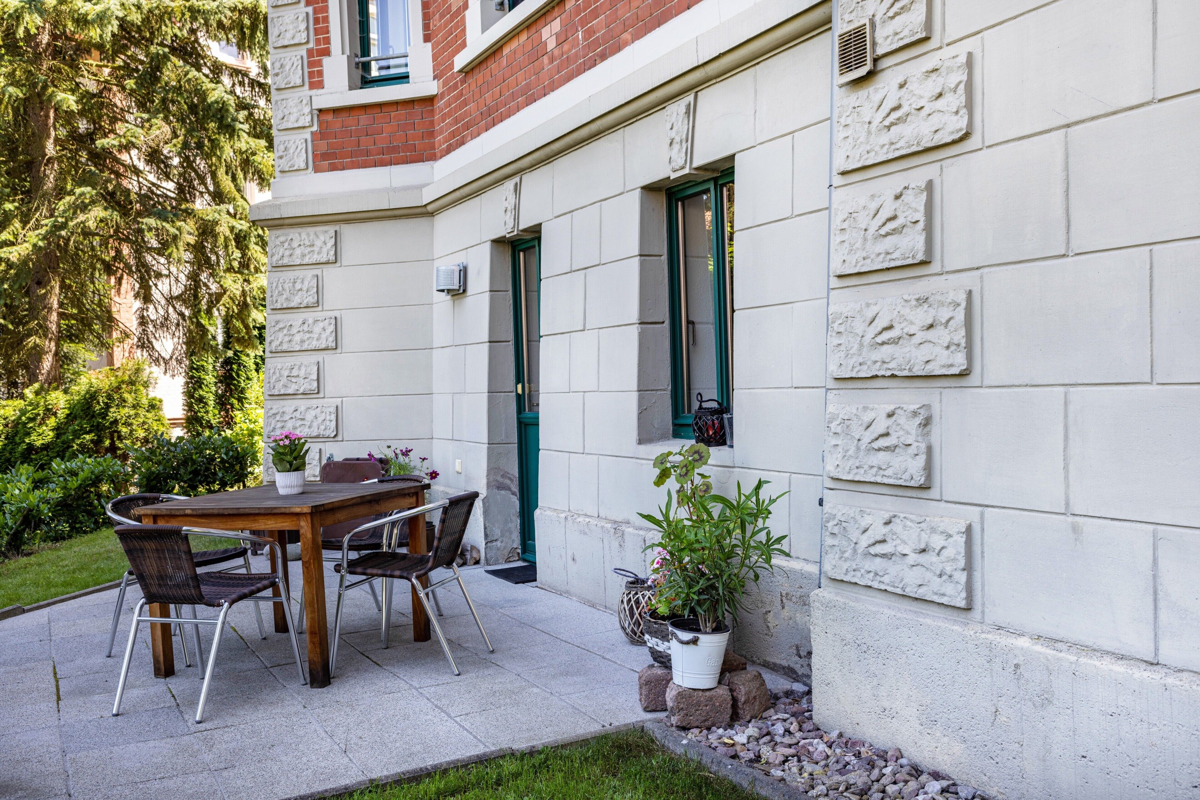 Eine Terrasse mit Holztisch und -stühlen, Blumentöpfen und einem Backsteinhaus mit dekorativem Mauerwerk.