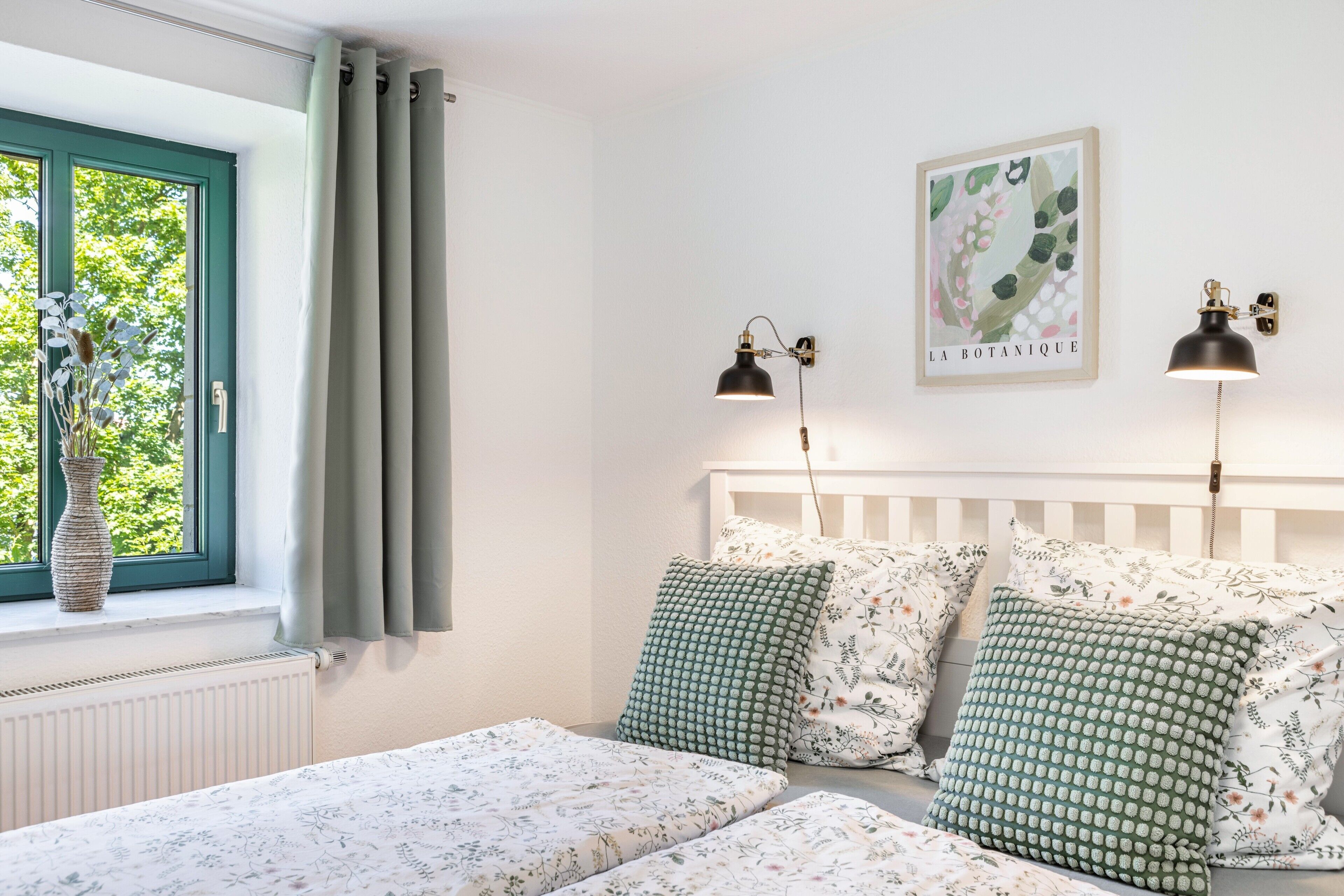 A bedroom with a white headboard, a window with curtains, a radiator, a bedside table with a vase, and a wall-mounted lamp.