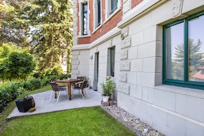 Une cour avec une table et des chaises, entourée de verdure et bordée d’un bâtiment à la façade en pierre et aux fenêtres apparentes.