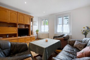 A living room with a leather sofa, a table with a tablecloth, wooden cabinets, a television, and a window with curtains. - Holiday Apartment 'Gästewohnung Am Schwanenteich' with Wi-Fi (Mittweida)