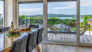 Een moderne eetruimte met een grote tafel en stoelen, een wijnfles en een glas. Buiten is er een balkon met uitzicht op een meer en groen.