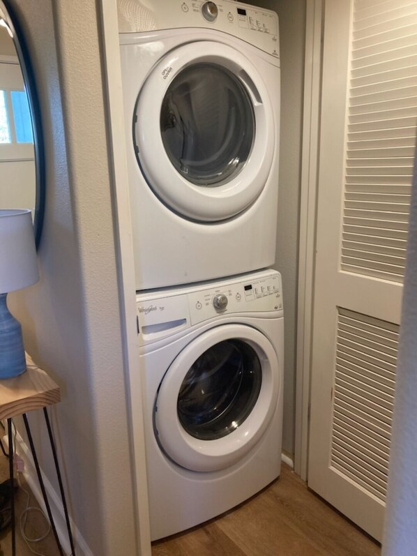 A compact, two-basin front-loading washer and dryer unit in a laundry room with a wooden floor and a white door with horizontal blinds. - Ko Olina Secluded Luxury: 3 Br, 2.5 Bath End Unit Townhome with Resort Amenities (Kapolei)