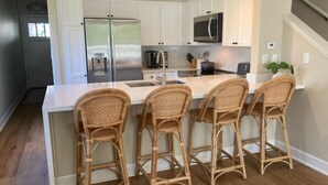 A kitchen with white cabinets, a stainless steel refrigerator, a microwave, and a sink. There are three wicker bar stools and a potted plant on the counter.