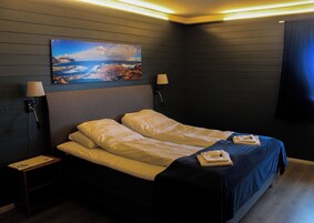 Una cama doble con sábanas blancas, dos lámparas de noche, un cuadro de un paisaje costero y una colcha azul oscuro.