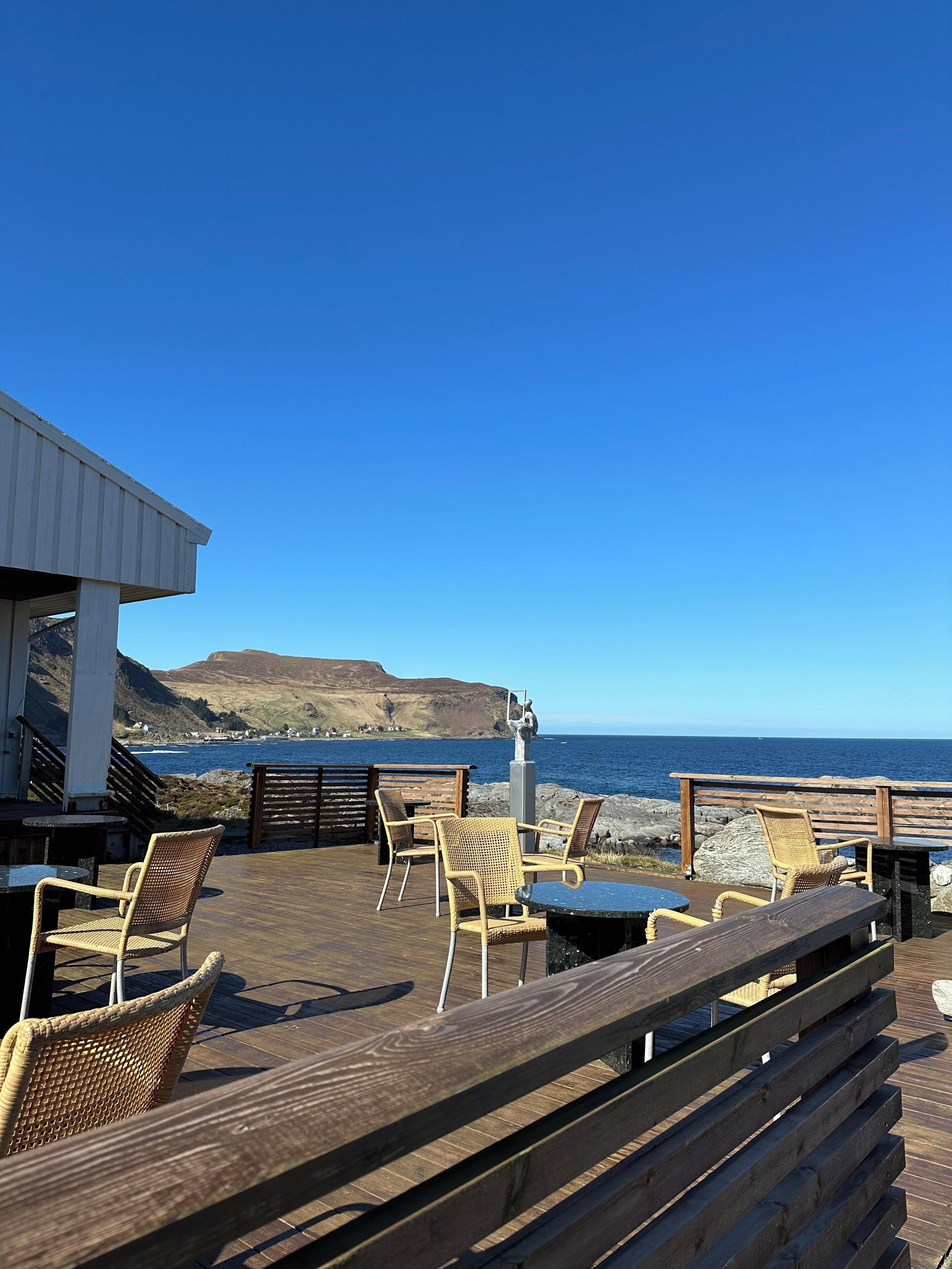 A wooden deck with wicker chairs and tables overlooking a coastal view.