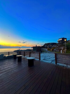 Una terraza de madera con bancos y mesas, con vistas a la costa durante el atardecer.