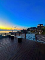 Una terraza de madera con bancos y mesas, con vistas a la costa durante el atardecer.