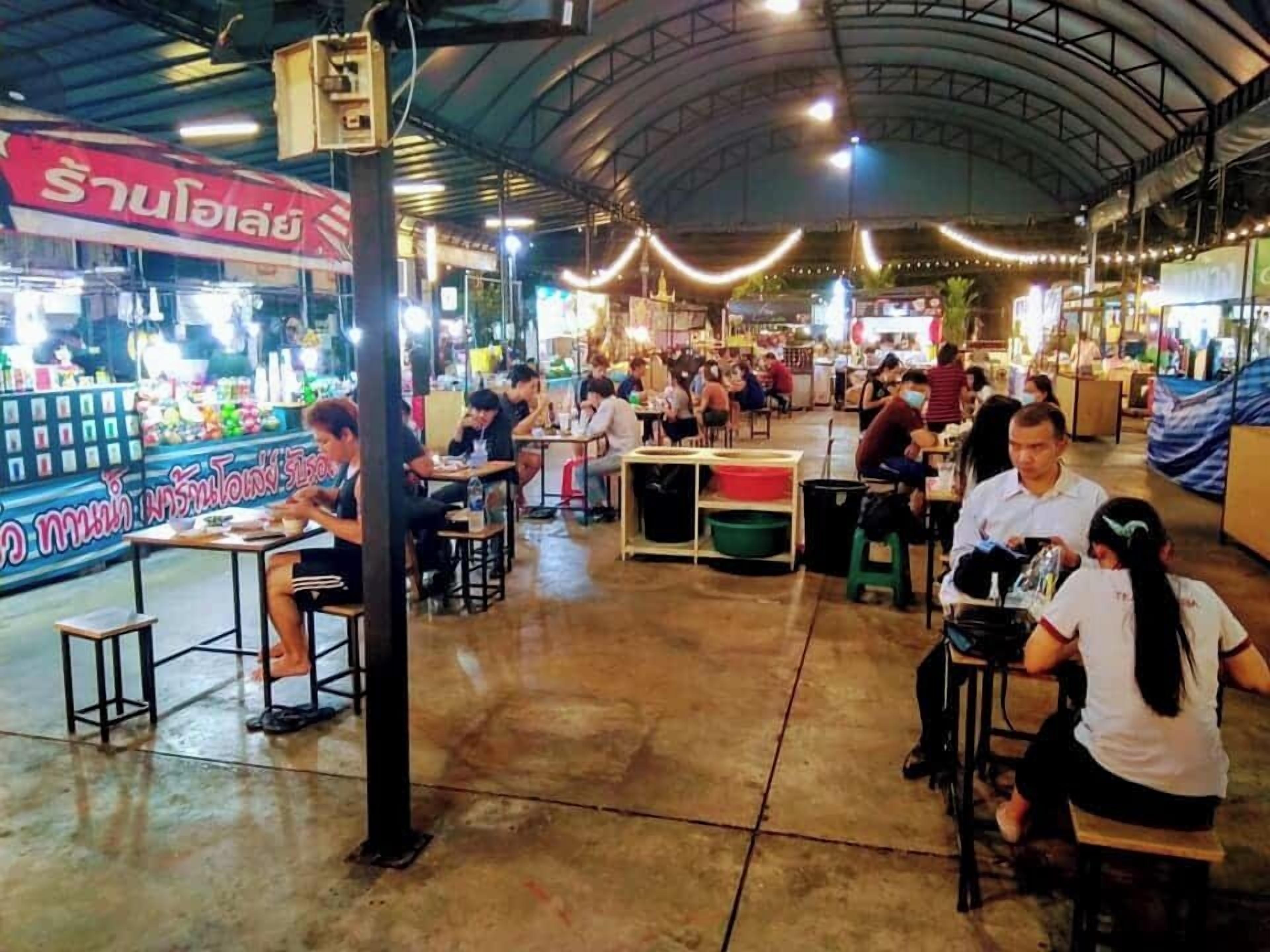 A bustling indoor market with tables and chairs for dining, a large sign with Thai text, and a high ceiling with lighting fixtures.