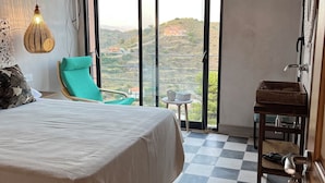 A bedroom with a large bed, a green chair, a small table with a cup, and a view of a hilly landscape through large windows.