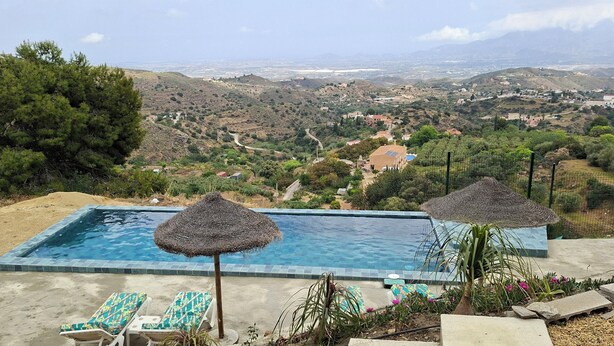 Une piscine à débordement avec deux parasols en chaume, des chaises longues et une vue sur un paysage vallonné.