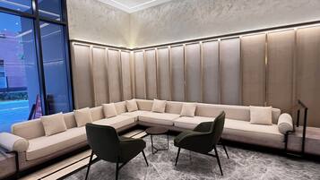 A modern lounge area with a large L-shaped sectional sofa, two armchairs, and a small round table. The room features a marble floor, a large window with a view of the outside, and a ceiling with recessed lighting.