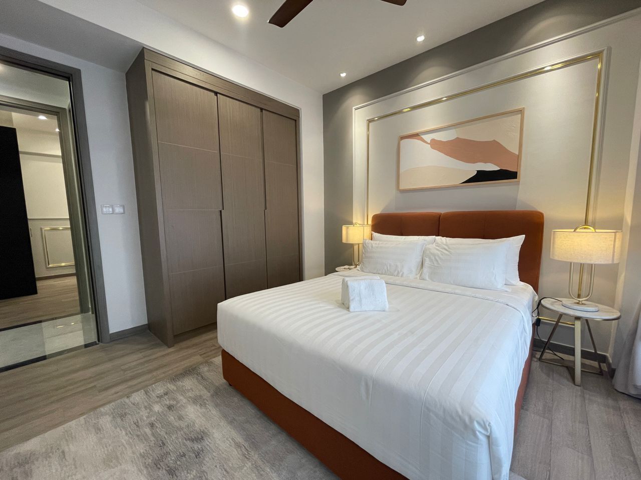 A modern hotel room with a large bed, a wardrobe, a bedside table with a lamp, and a framed artwork on the wall.