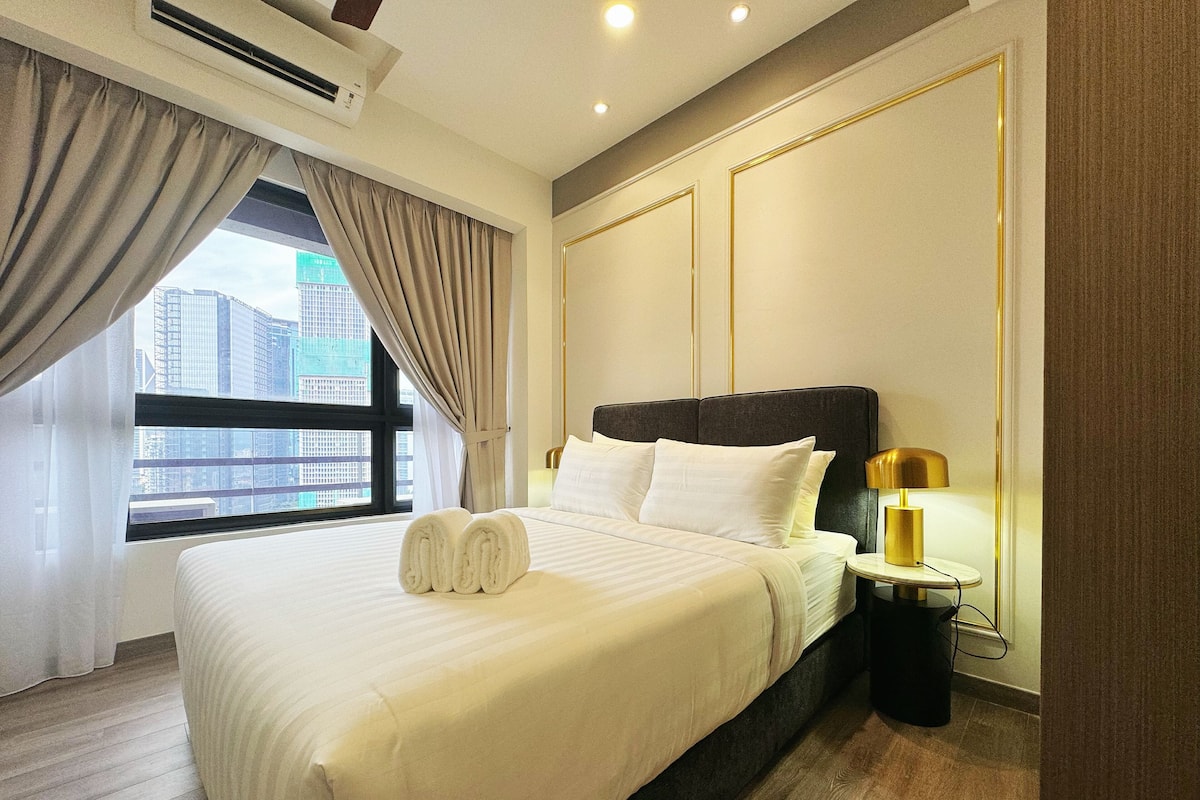 Habitación de hotel con una cama grande, una mesita de noche, una lámpara y vistas a la ciudad a través de una ventana.