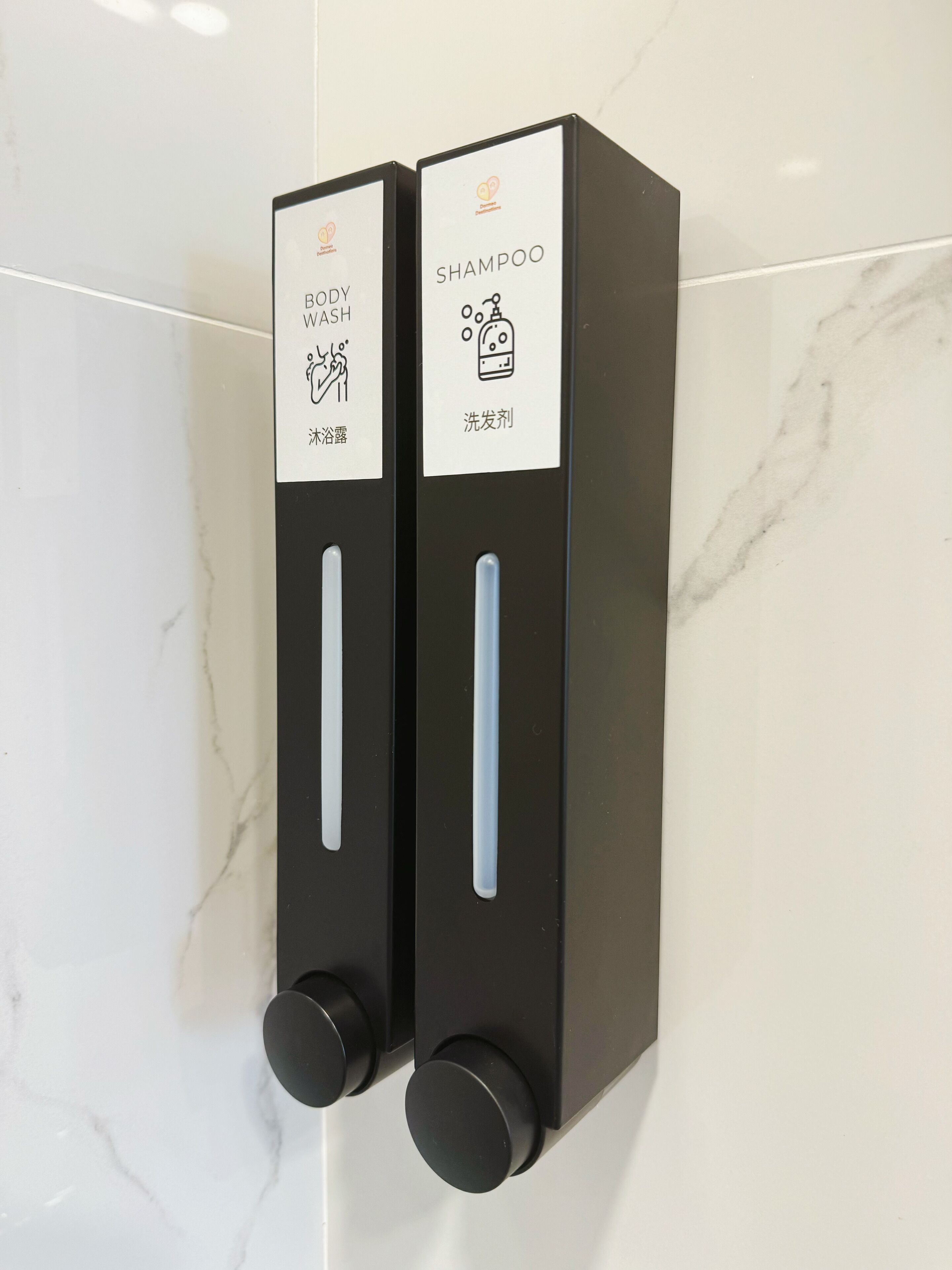 Two black dispensers with labels for body wash and shampoo, mounted on a white tiled wall.